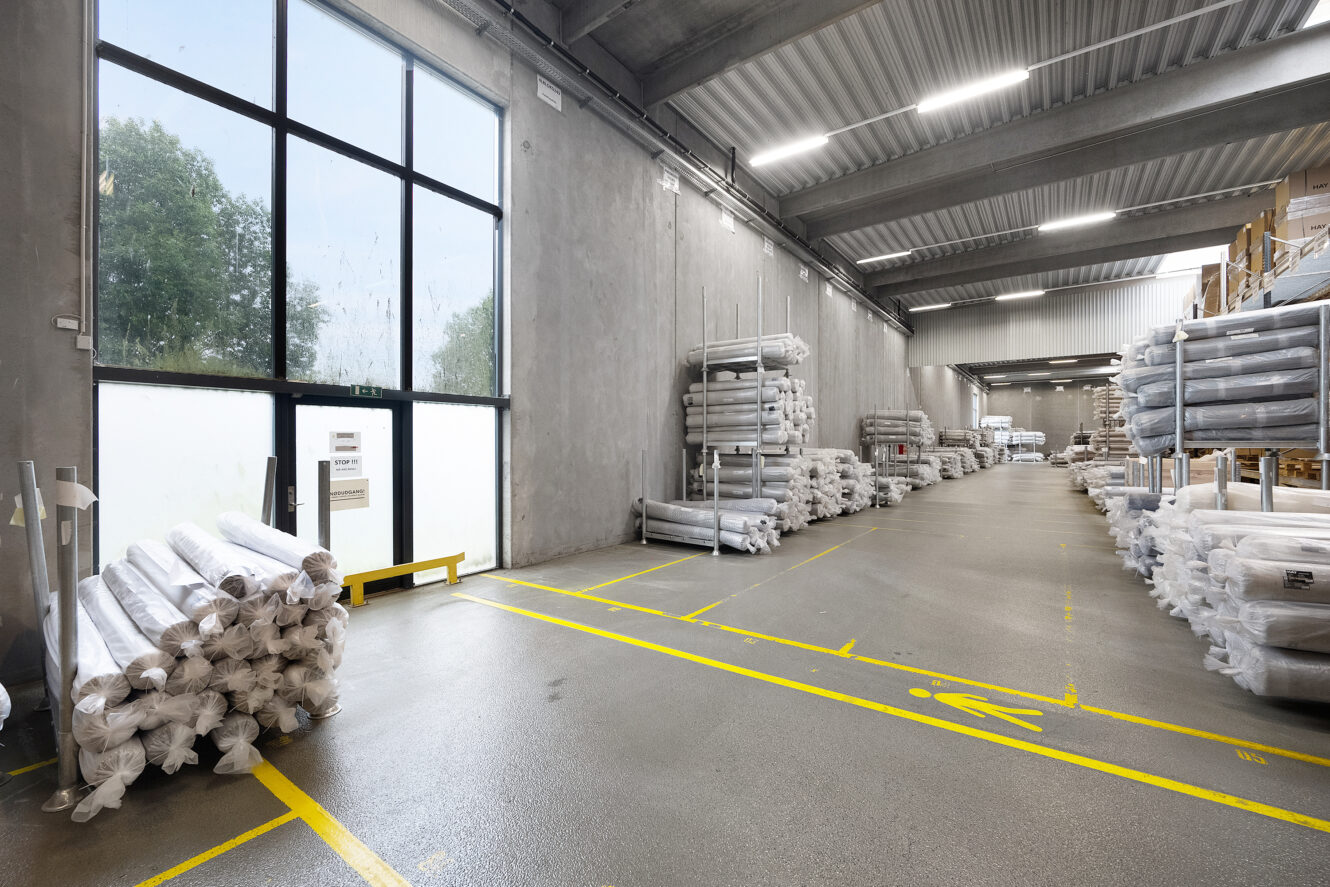 Et lagerinteriør med store vinduer, betonvægge og stabler af valsede materialer på metalreoler langs begge sider af en bred gang.