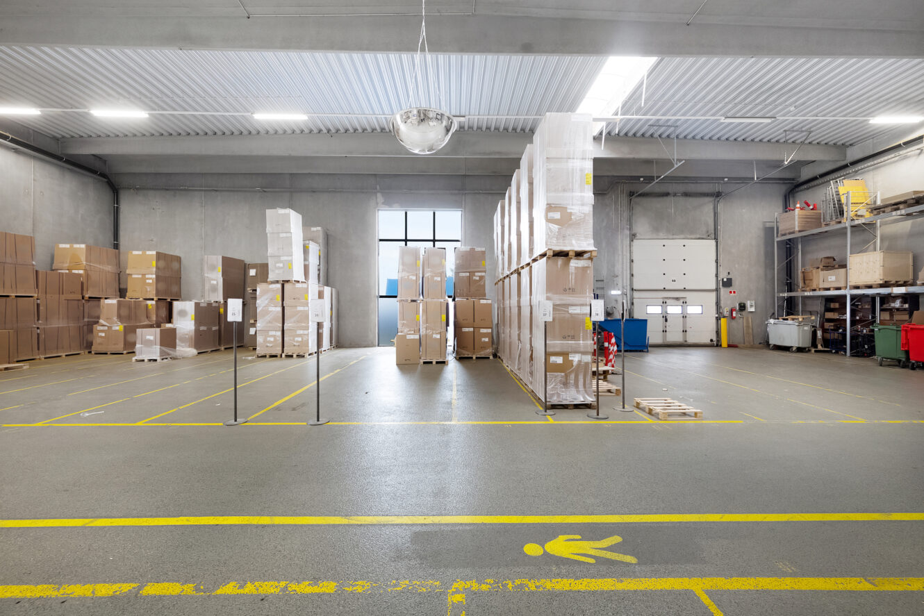 Stort lagerinteriør med stablede paller med kasser, gule sikkerhedslinjer på gulvet og betonvægge under skarpe loftslamper.