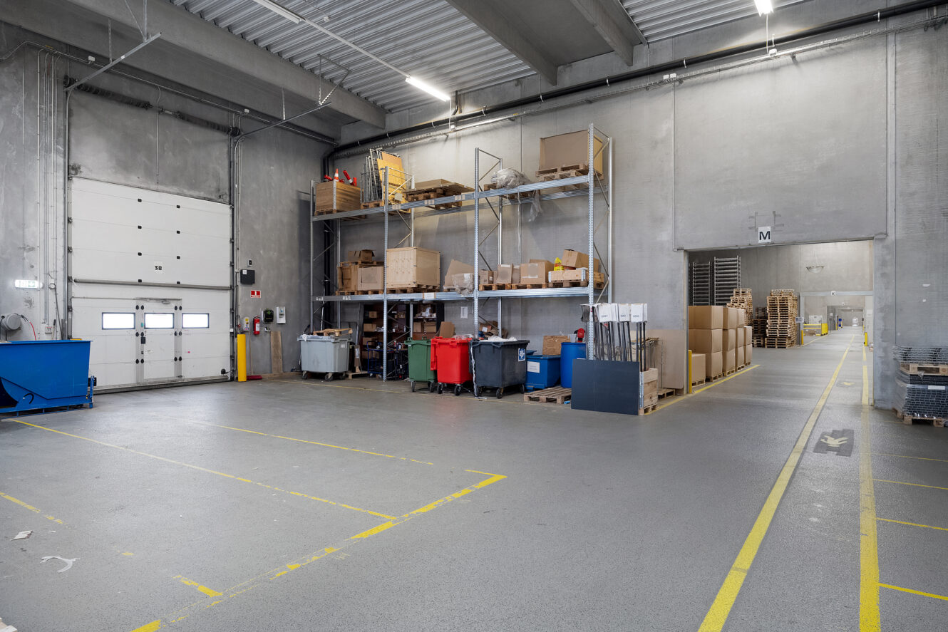 Et rent lager med stablede hylder med kasser og forskellige varer, gule sikkerhedslinjer på gulvet og en lukket dør til læsserampen til venstre.