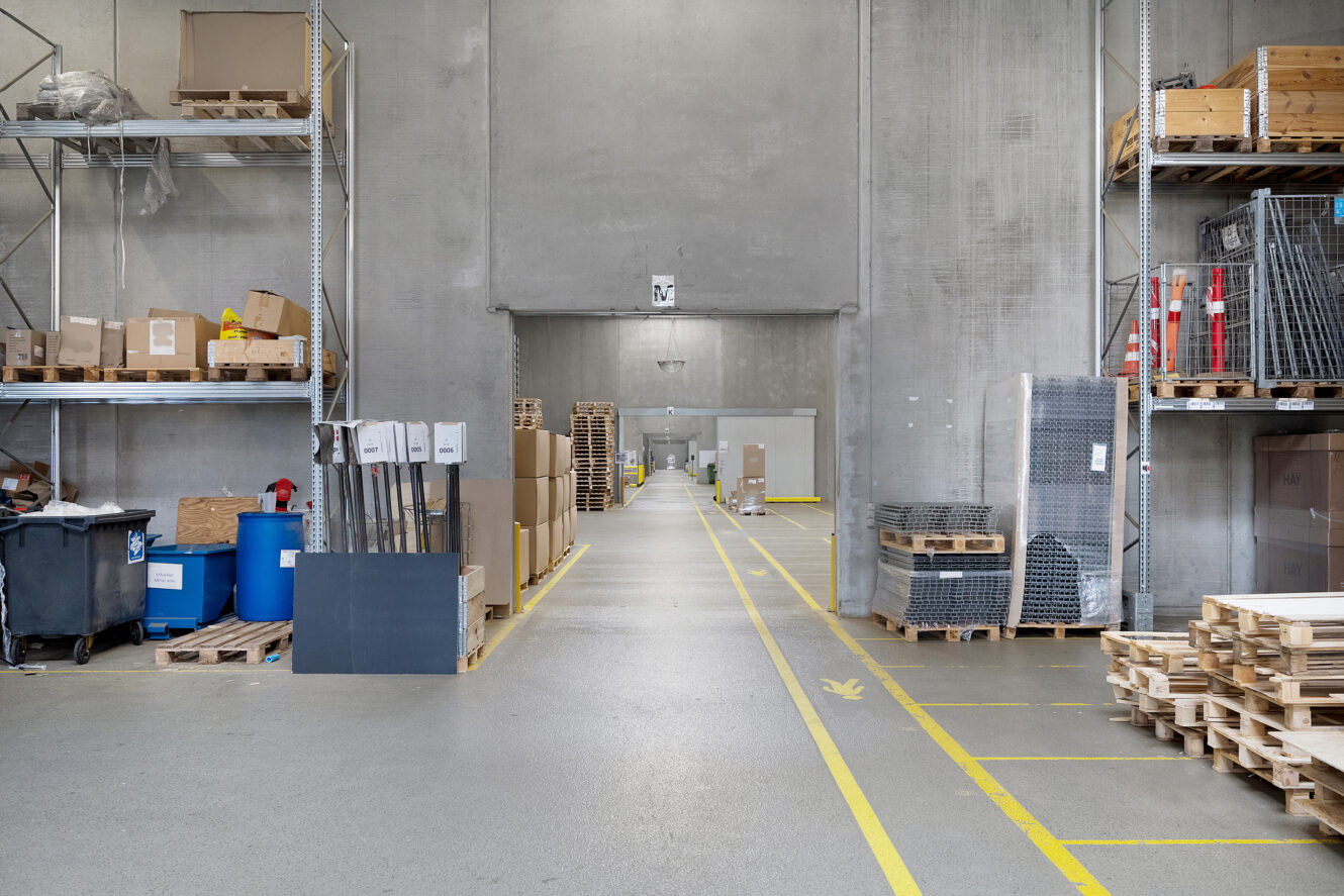 Lagerinteriør med hylder, paller og papkasser organiseret langs en bred gang; betonvægge og industriel belysning er synlige.