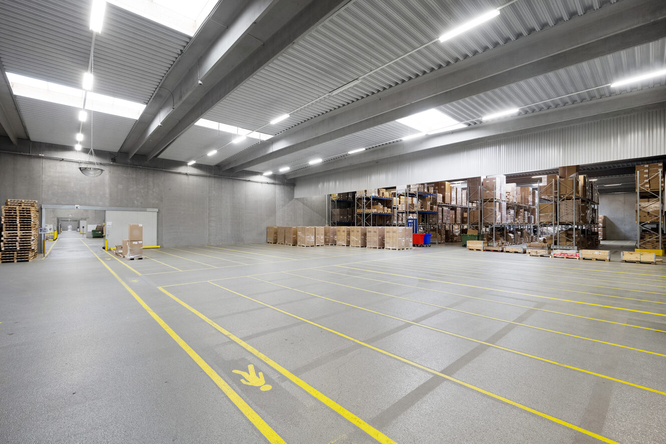 Rummelig lagerbygning med højt til loftet, reoler fyldt med kasser, træpaller og gule streger, der markerer sektioner på gulvet.