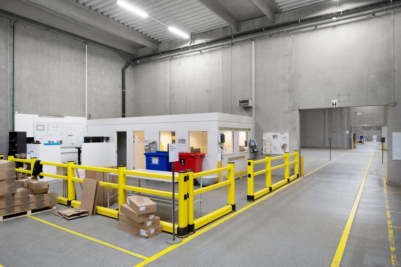 En lagerbygning med betonvægge, gule sikkerhedsbarrierer, et lille kontor og stabler af papkasser på gulvet.