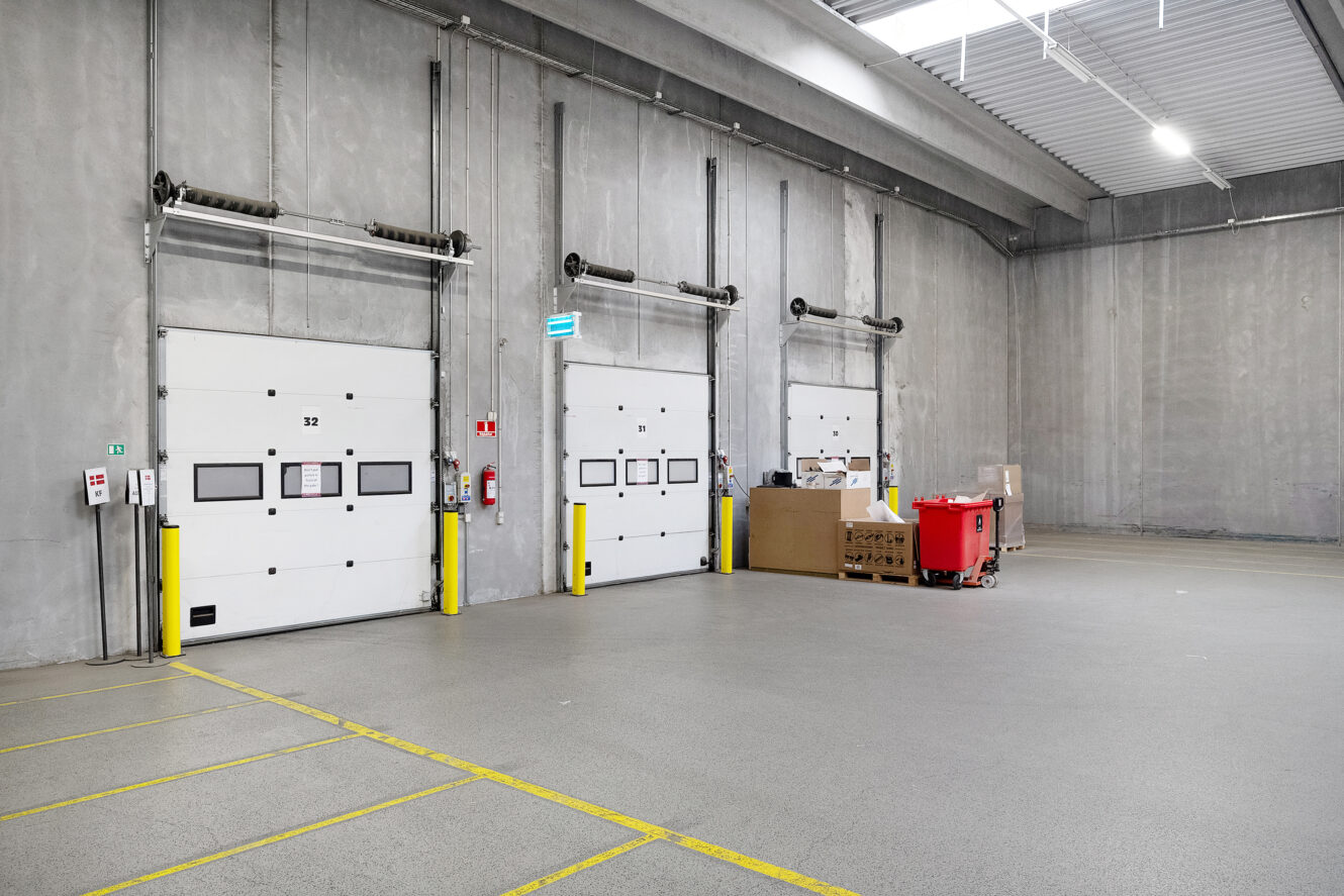 Interiør af en tom lagerhal med tre store læsseramper, industriel belysning og nogle paller og beholdere mod væggen.