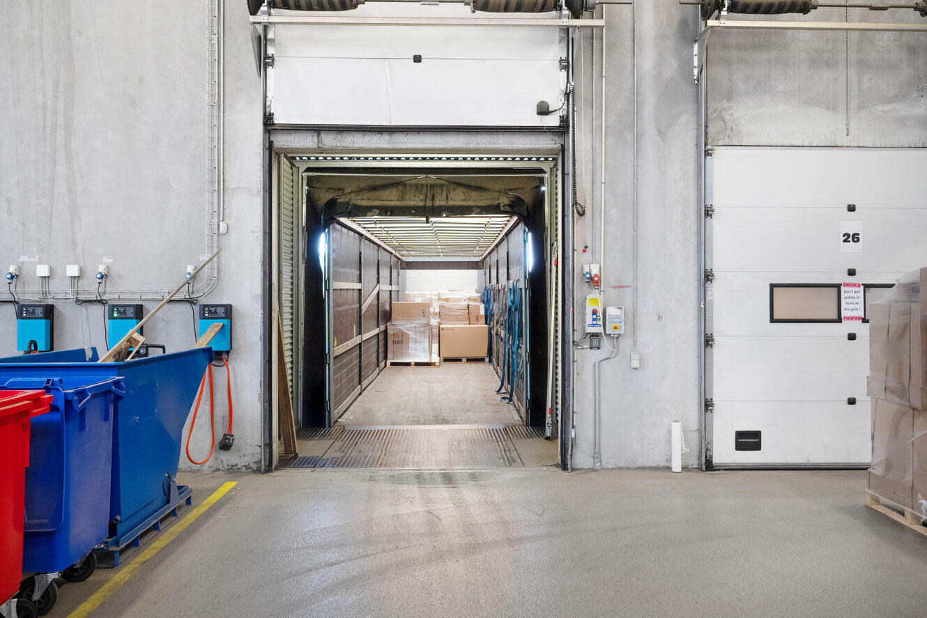 Læssekaj med en åben lastbil, der bakker op, palleterede varer indeni og beholdere og kasser i nærheden i et lagermiljø.