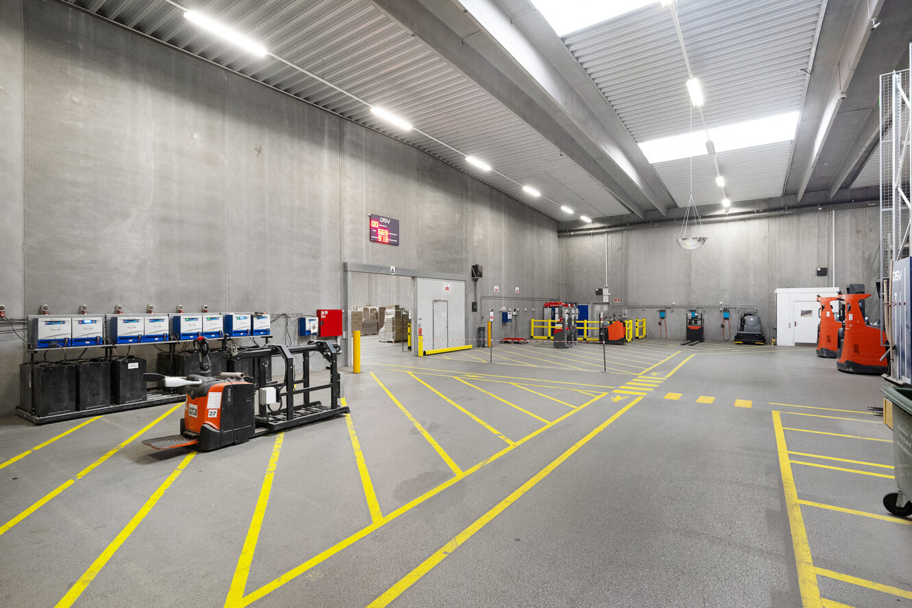 Et rummeligt industrilager med højt til loftet, tomme palleløftere, batteriladestationer og gule sikkerhedslinjer, der markerer gulvet.