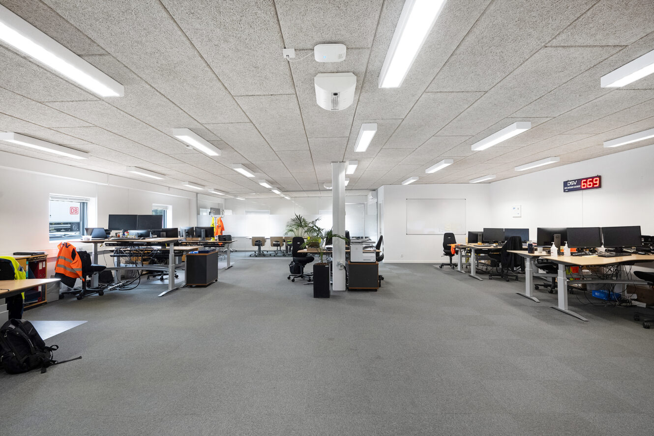 Et moderne åbent kontor med flere skriveborde, computere, stole og jakker med høj synlighed, med kraftig belysning og et digitalt vægur, der viser 6:59.