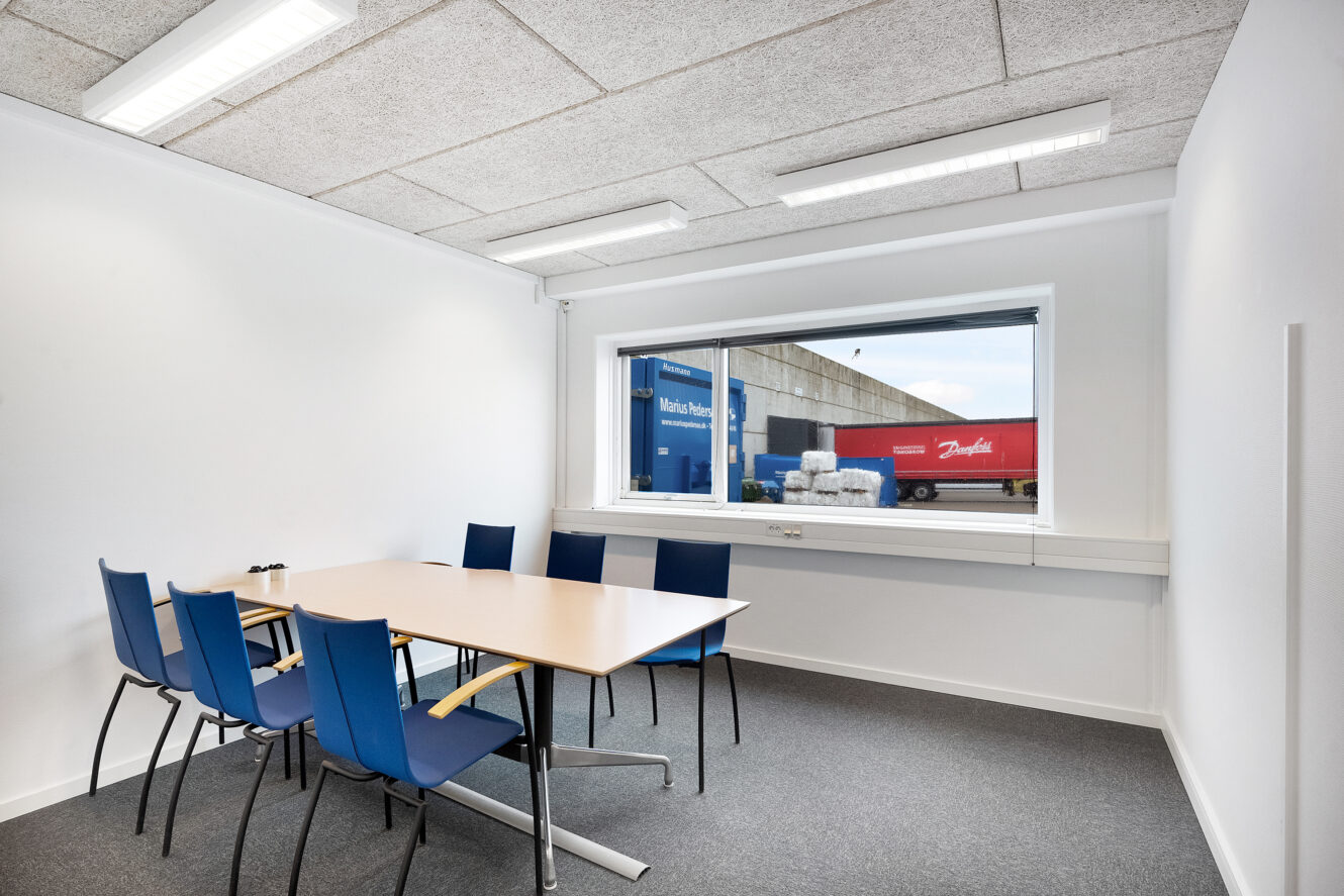 Et lille mødelokale med et bord, seks blå stole og et stort vindue med udsigt til en industrigård med lastbiler og paller.