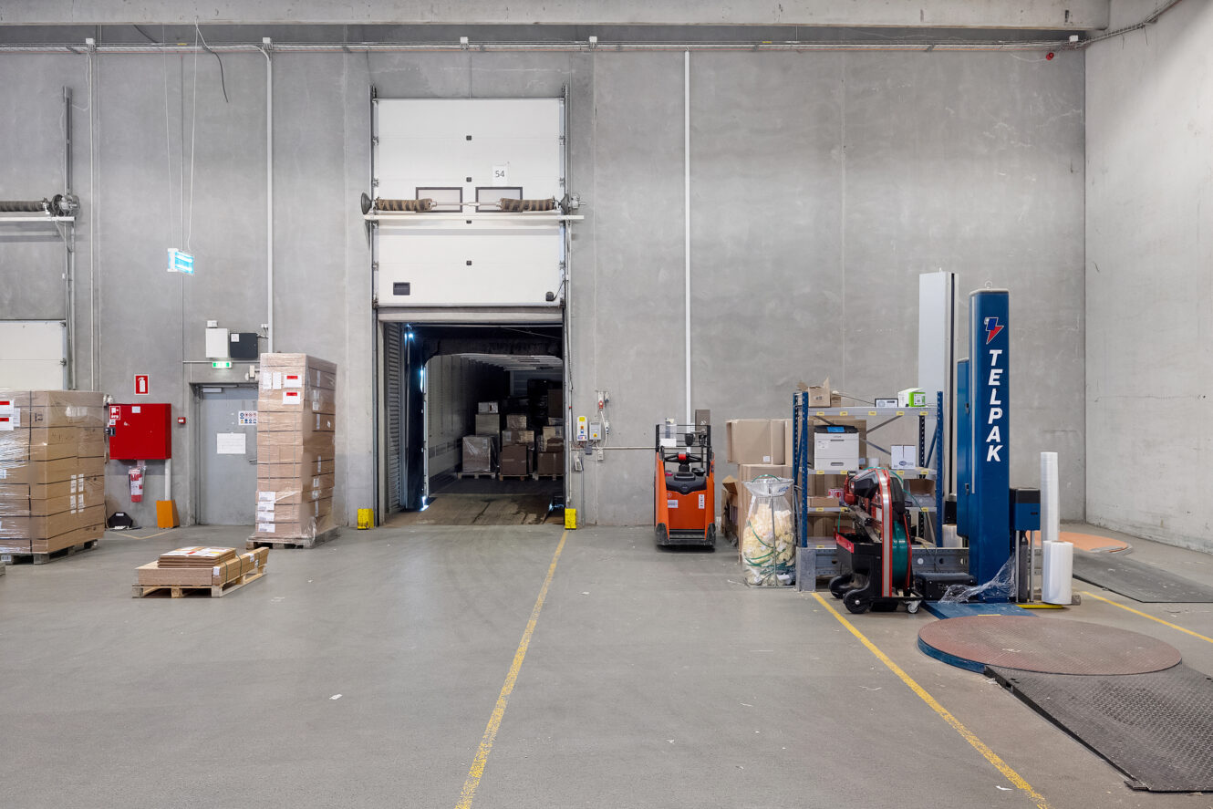 En læsserampe på et lager med paller med kasser, en gaffeltruck og emballeringsudstyr; en lastbil er delvist læsset ved den åbne dør.