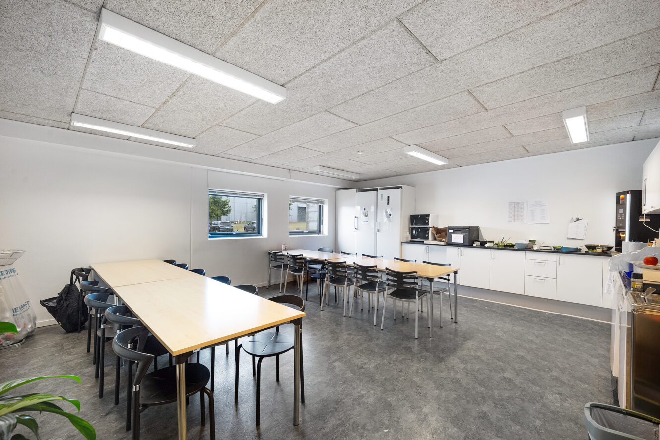 Et rent, moderne pauserum med to lange borde, sorte stole, hvide skabe, køkkenmaskiner og et vindue med udsigt til det fri.