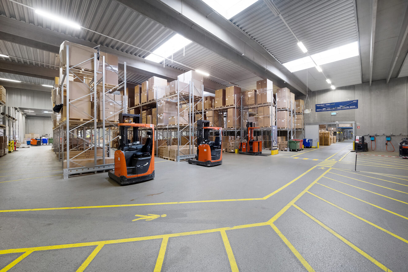 Rummelig lagerbygning med hylder med stablede kasser, flere orange gaffeltrucks, betonvægge og markerede gule linjer på gulvet.