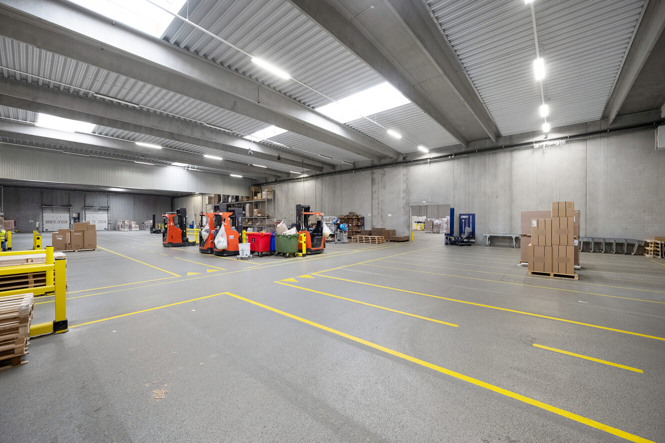 Stort, rent lagerinteriør med højt til loftet, gaffeltrucks, stabler af papkasser og gule streger, der markerer lager- og trafikområder på det grå gulv.