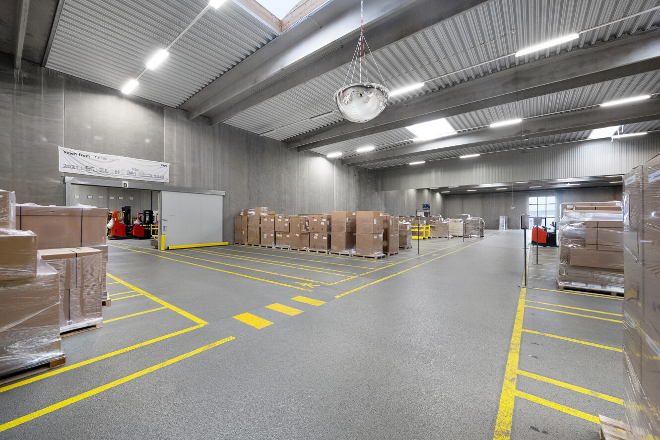 Et rummeligt lagerinteriør med stablede paller af papkasser, gaffeltrucks i baggrunden og gule markeringer på det grå gulv.