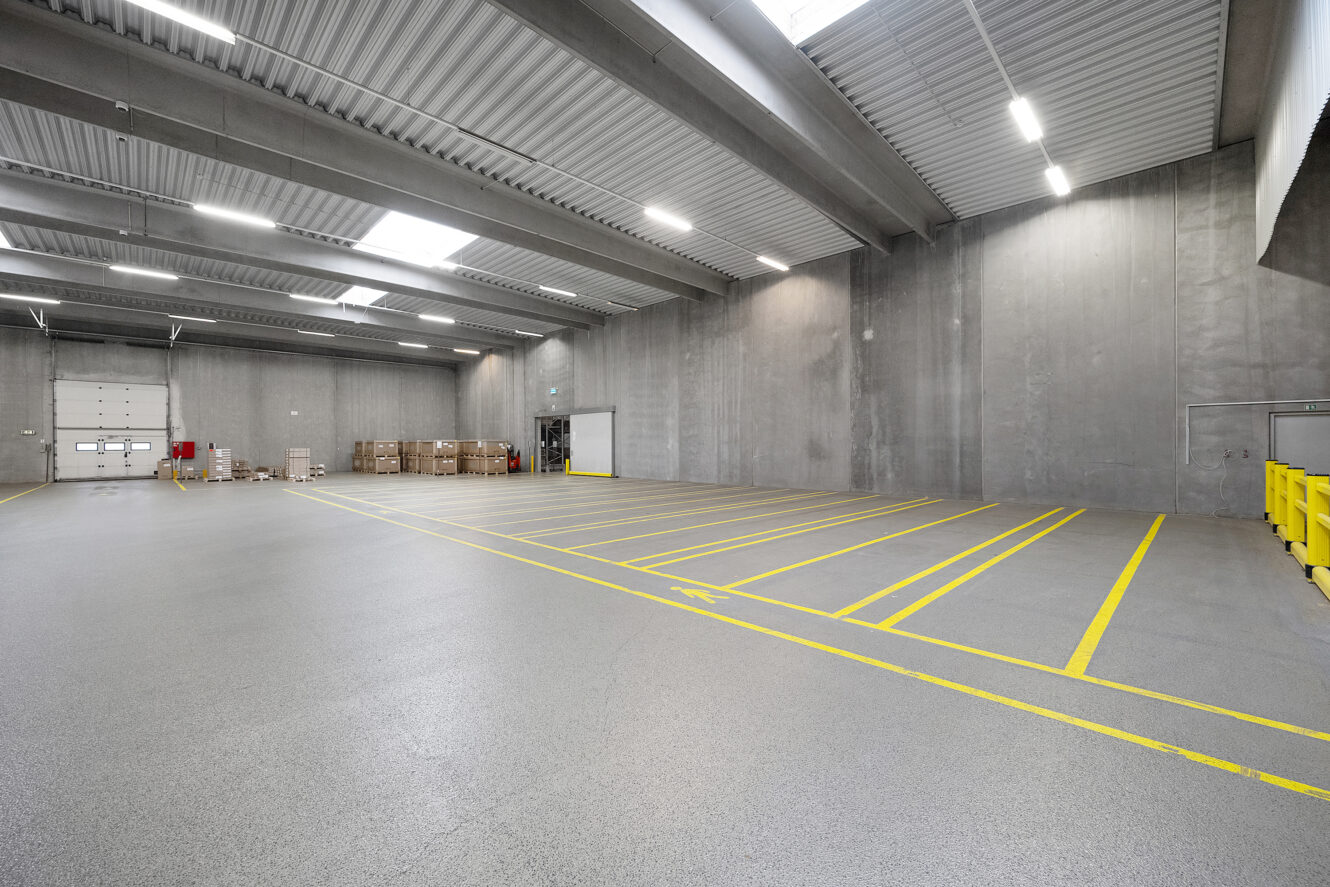 Stor tom lagerbygning med højt til loftet, betonvægge, afmærkede parkeringspladser, paller og kasser stablet nær en læsserampe.