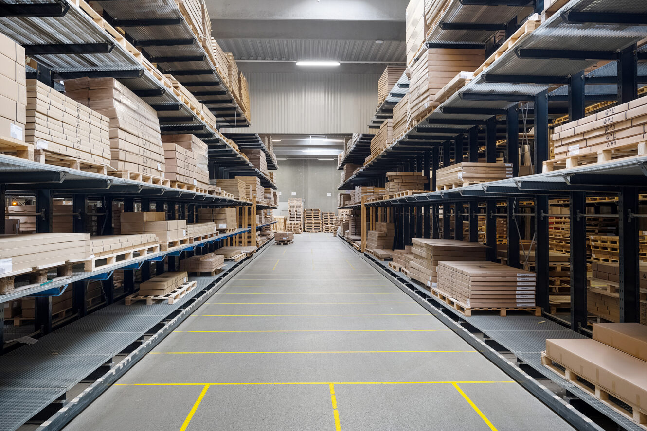 Lageret er indrettet med høje hylder fyldt med papkasser og træpaller og en bred gang i midten.