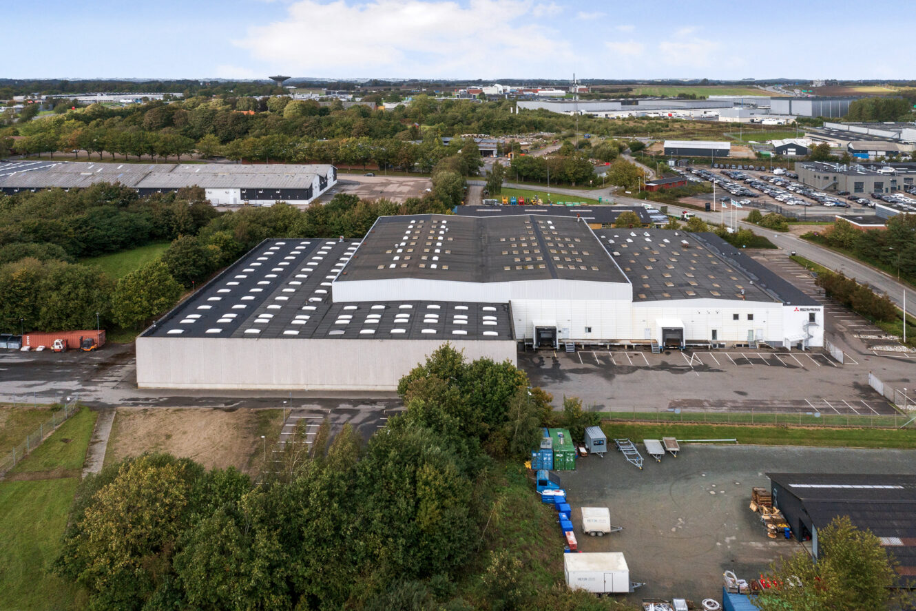 Et stort industrilager med et hvidt og gråt ydre omgivet af træer og parkeringspladser med flere synlige læsseramper.