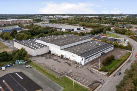 Luftfoto af et stort industrilager med parkeringspladser, omgivet af træer og nærliggende veje i et erhvervsområde.