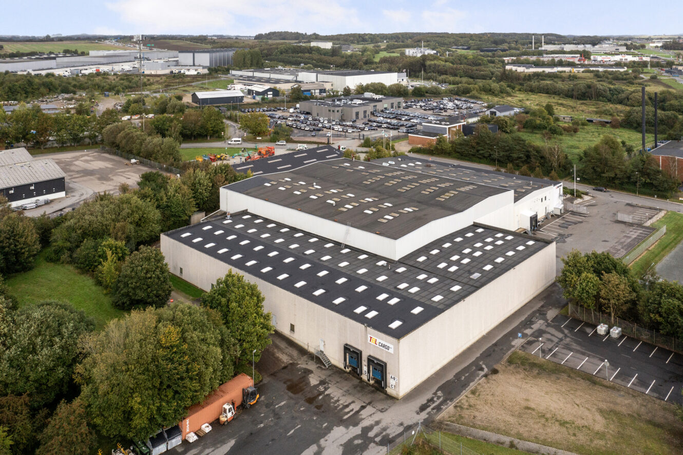 Luftfoto af et stort industrilager omgivet af træer, parkeringspladser og nærliggende industribygninger.