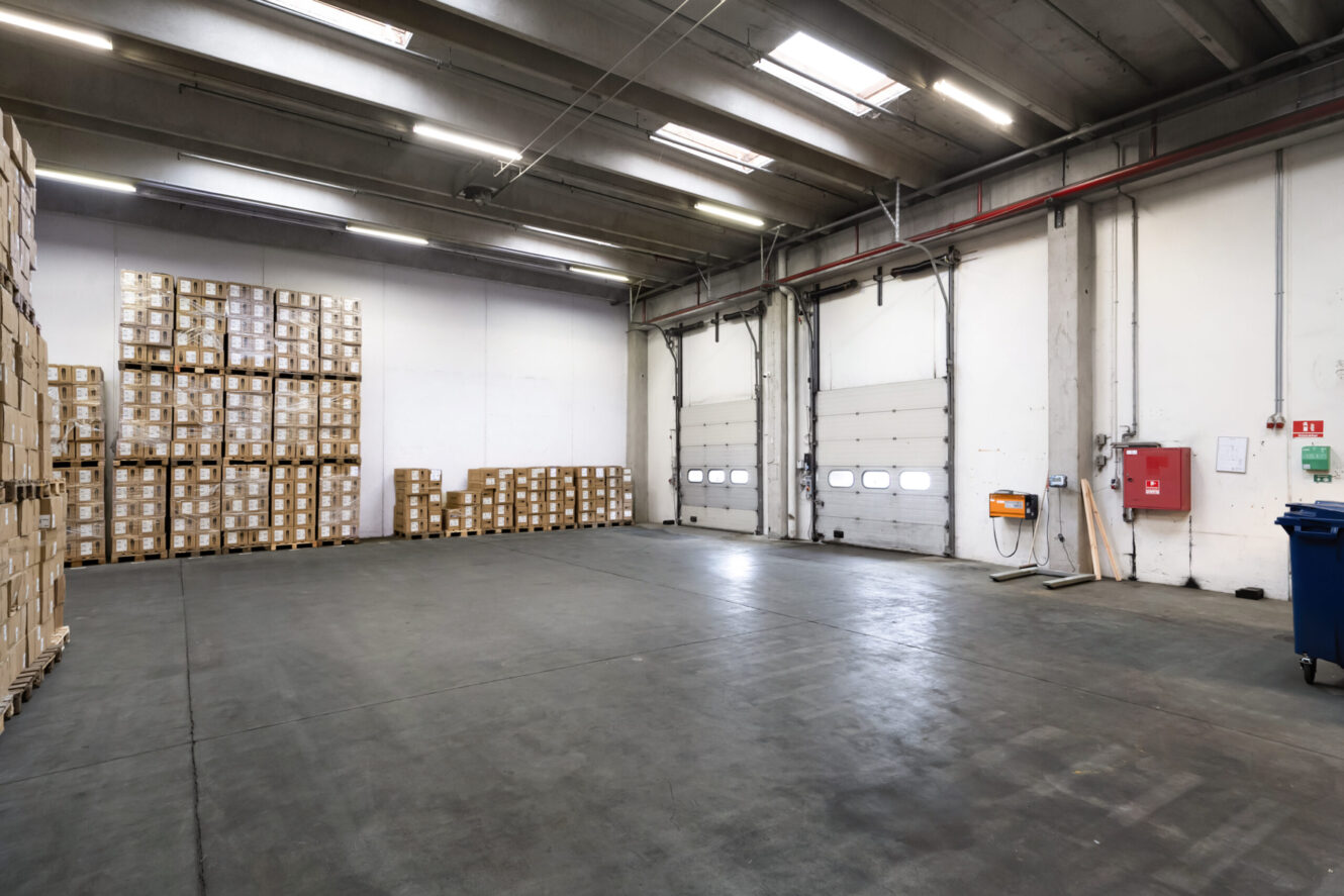 Stor, tom lagerhal med betongulv, højt til loftet, stablede papkasser langs venstre væg og tre lukkede læsseramper til højre.