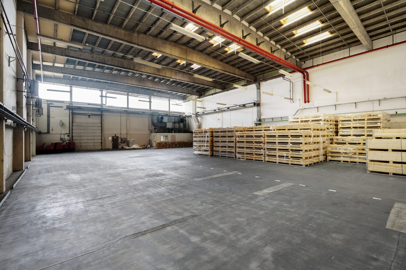 Rummelig lagerbygning med stabler af træpaller i højre side og store vinduer, der lukker naturligt lys ind.