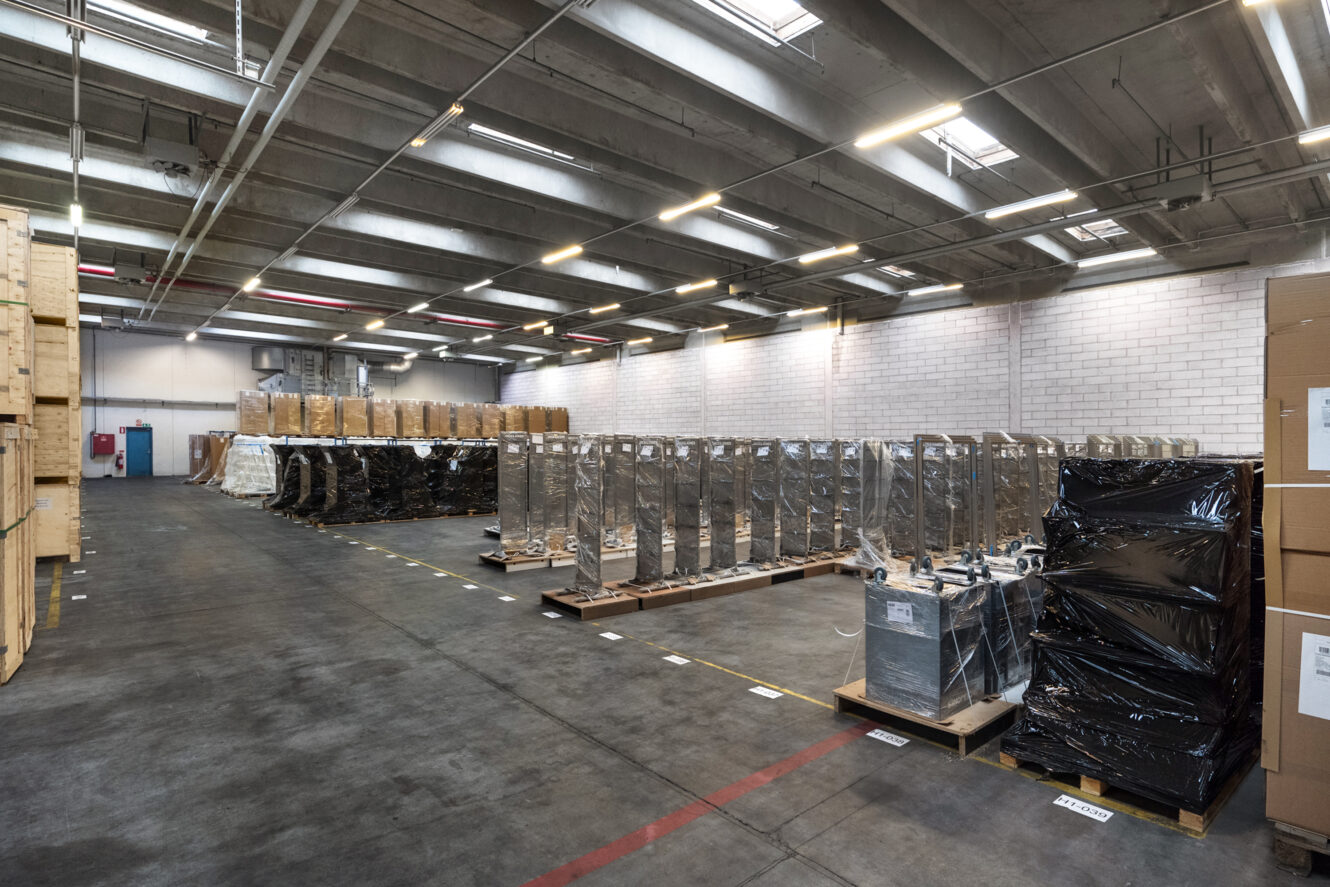 Stort lagerinteriør med stablede paller med varer, nogle pakket ind i plastik, under skarpe loftslamper og ovenlys.