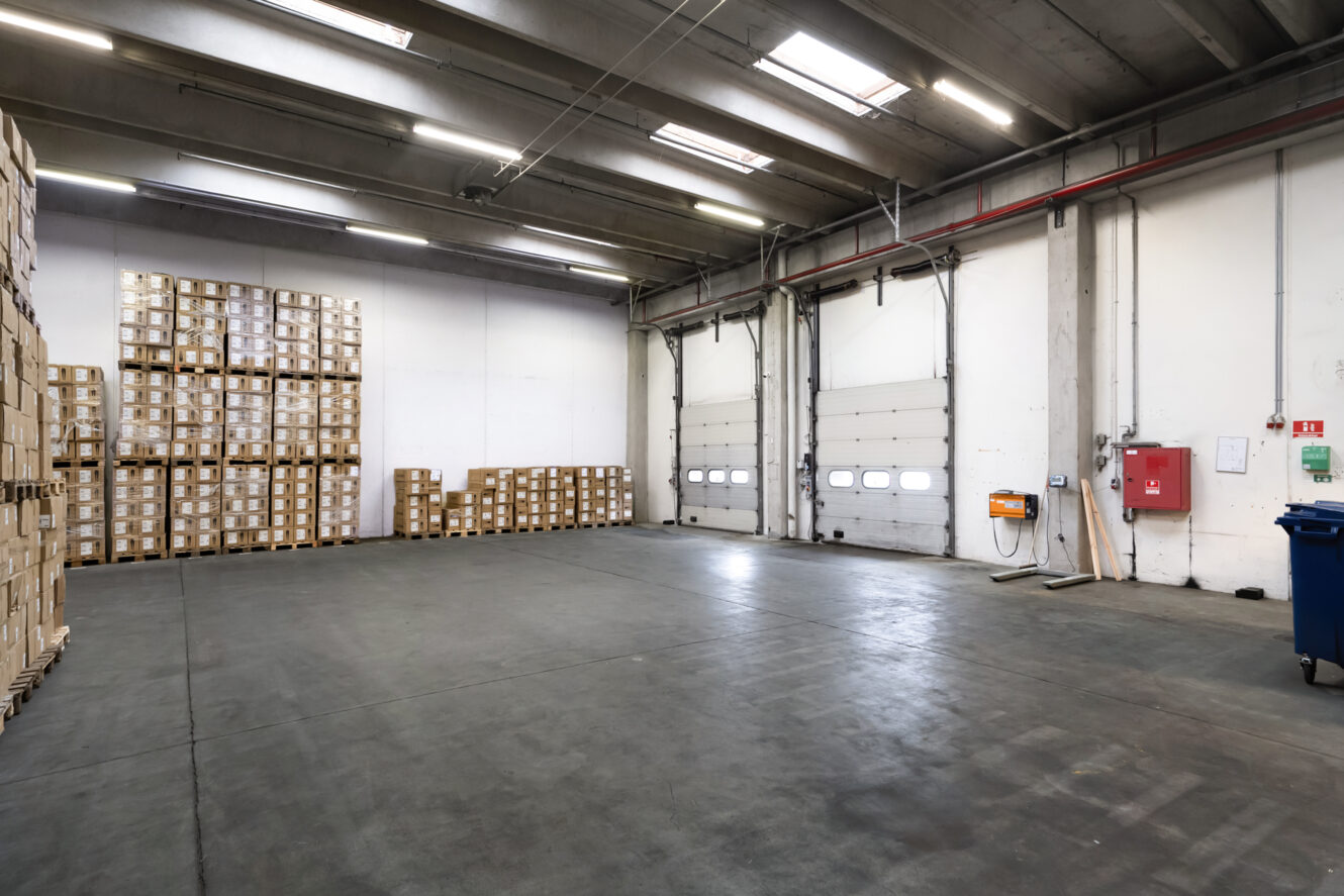 Interiør i en stor lagerhal med stablede papkasser langs venstre væg, betongulv, højt til loftet og lukkede porte til læsserampen til højre.