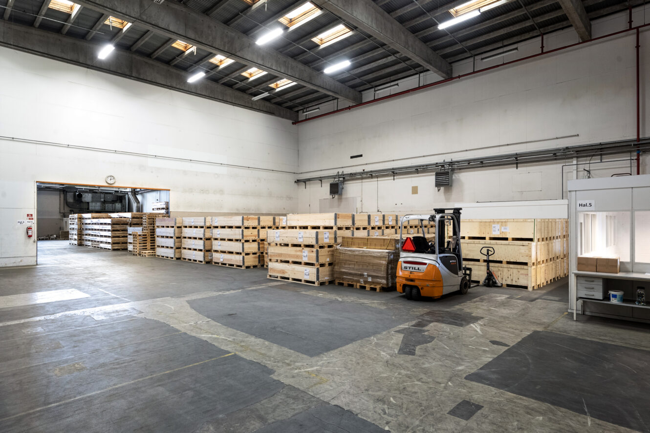 Et lagerinteriør med stablede trækasser, en orange gaffeltruck og industriel belysning over hovedet.