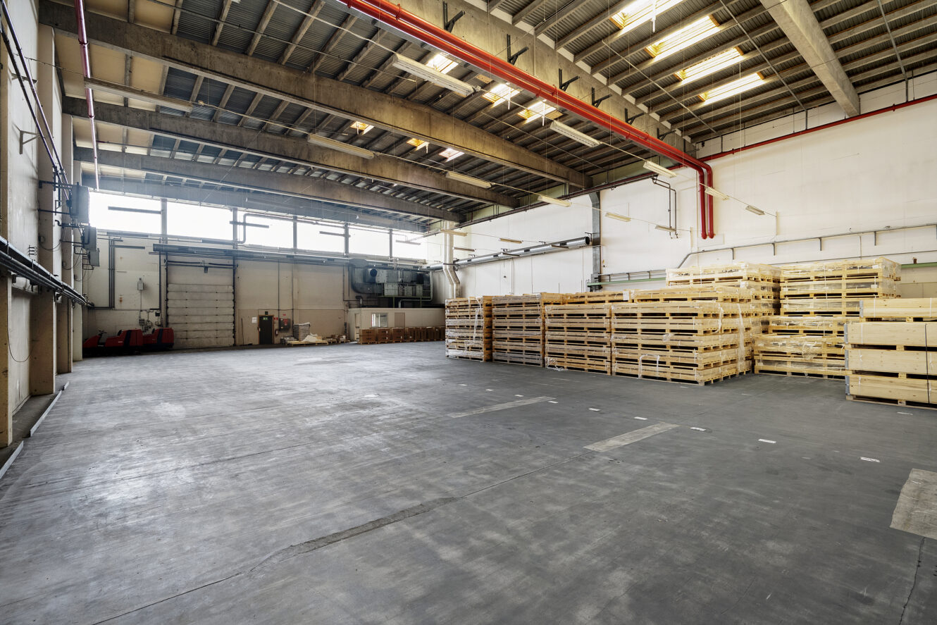 Rummelig industriel lagerbygning med højt til loftet, store vinduer, stablede trækasser på den ene side og betongulv.