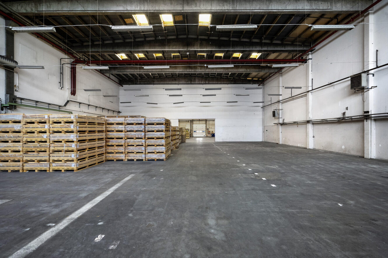 Rummeligt, tomt lagerinteriør med højt til loftet, stablede træpaller til venstre og betongulv.