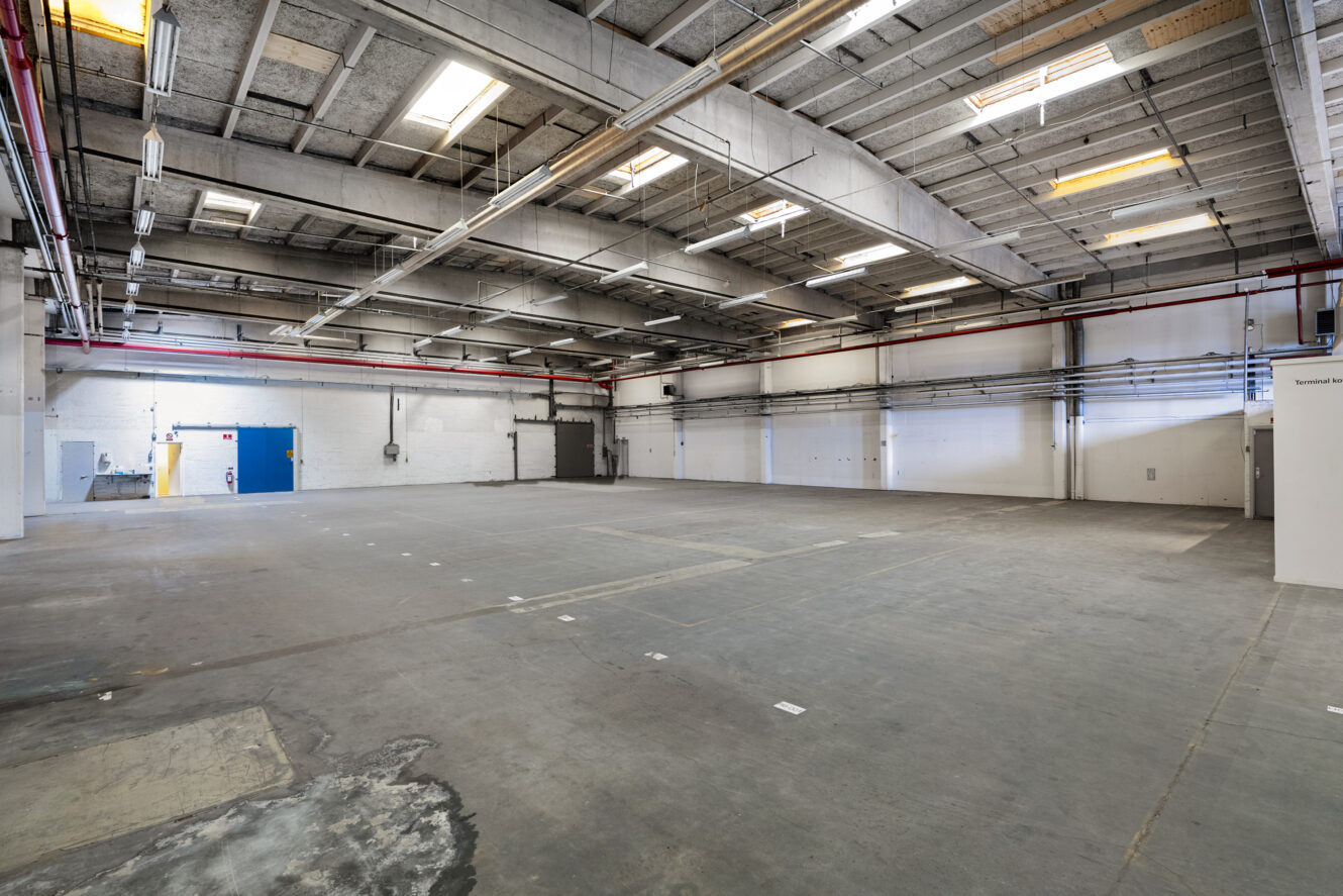 Stort tomt industrilager med betongulv, højt til loftet, synlige rør og fluorescerende belysning. Blå og grå døre er synlige langs bagvæggen.