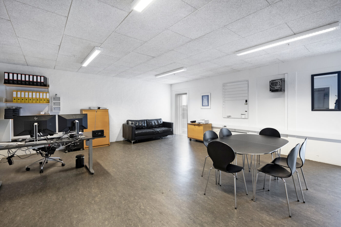 Et moderne kontor med skriveborde og computere til venstre, en sort sofa mod bagvæggen og et rundt bord med stole til højre.