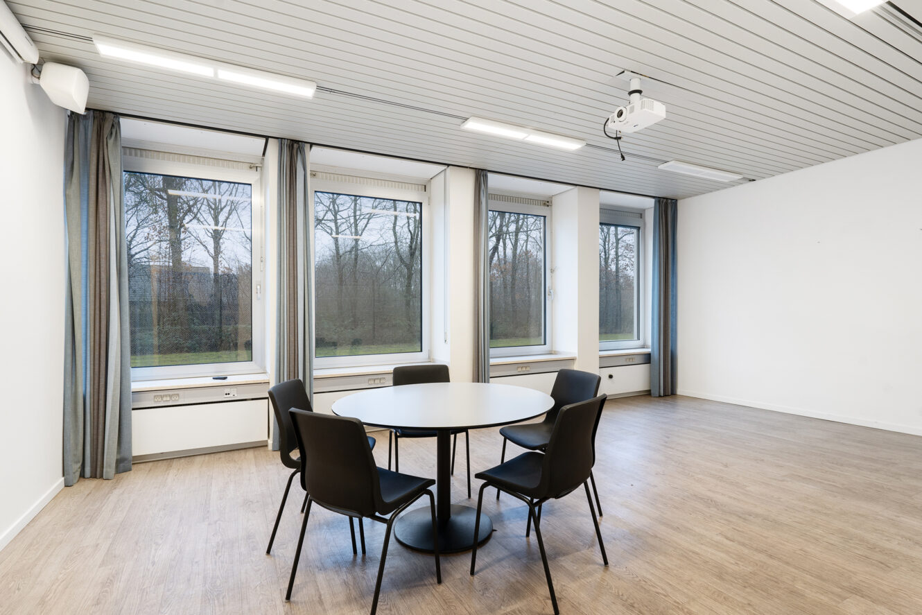 Et rundt bord med fem stole er placeret i et minimalistisk mødelokale med store vinduer, trægulv, hvide vægge og en loftsmonteret projektor.