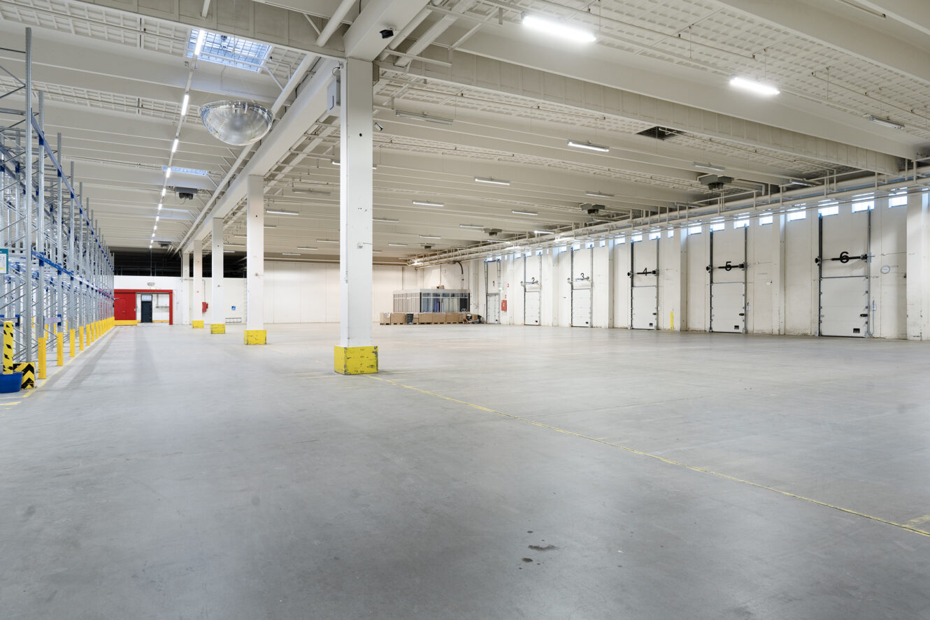 Rummeligt, tomt lagerinteriør med betongulv, højt til loftet, reoler til venstre og læsserampeporte på højre væg.