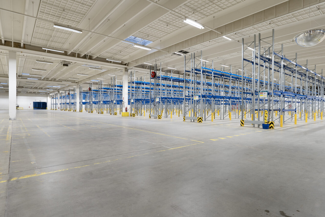 Stort, tomt lagerinteriør med høje metalreoler og kraftig ovenlys. Gulvet er markeret med gule linjer af hensyn til organisation og sikkerhed.