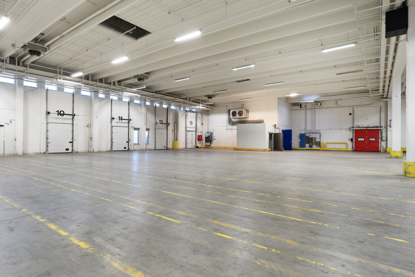 Rummelig, tom lagerhal med højt til loftet, porte til læsseramper, ovenlys og markerede linjer på betongulvet.