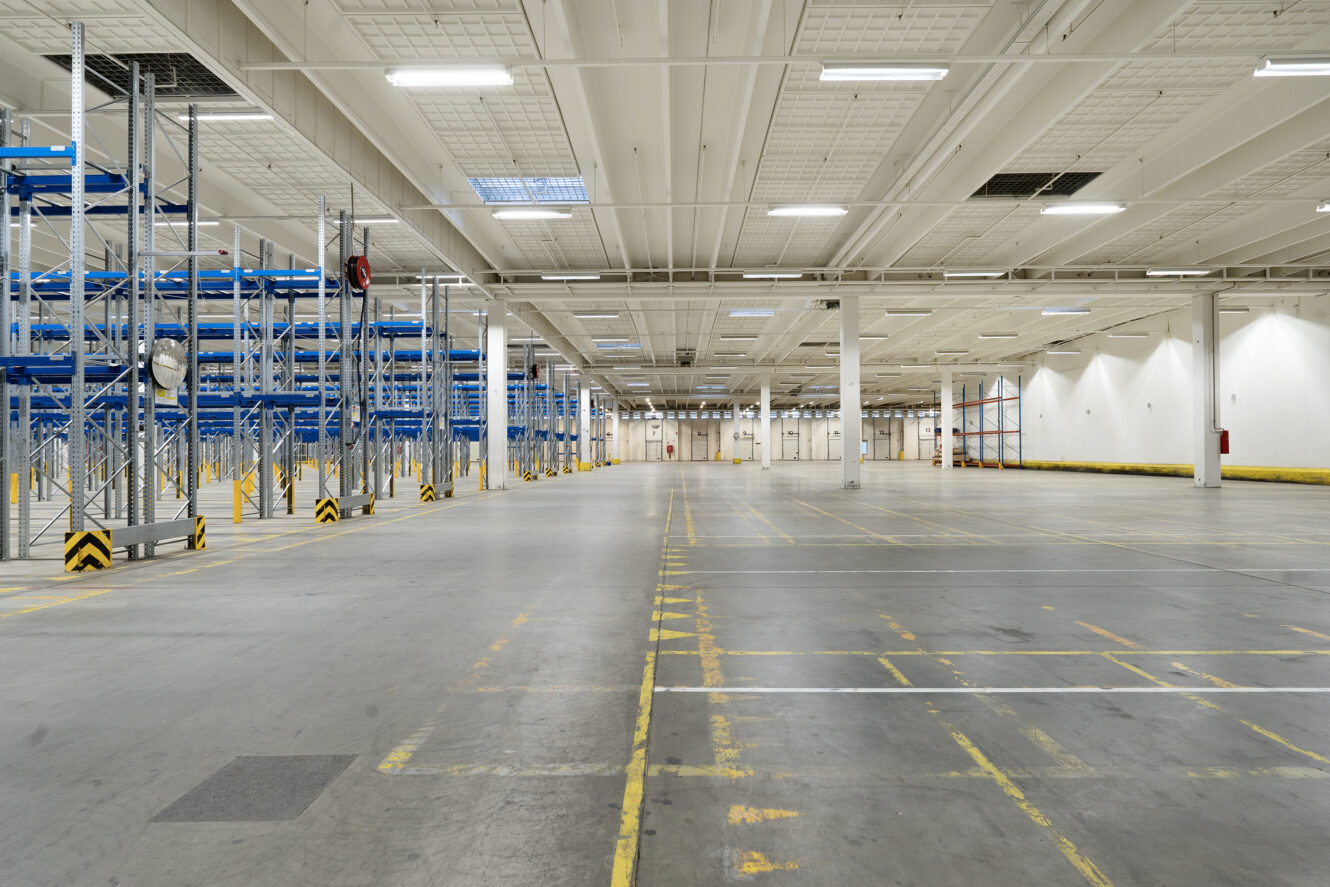 Bredt billede af en tom lagerhal med højt til loftet, metalreoler til venstre, gule sikkerhedslinjer på gulvet og kraftig ovenlys.