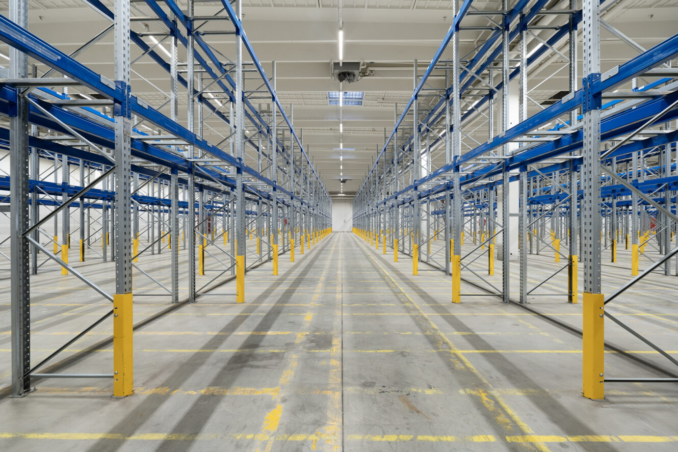 Tomt lagerinteriør med rækker af høje metalreoler, blå bjælker og gule sikkerhedsafskærmninger under kraftig ovenlys.