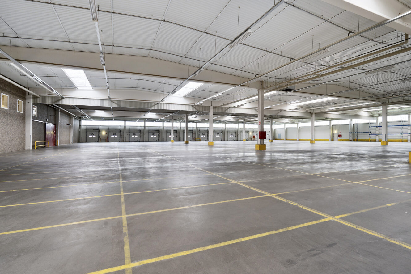 Stor tom lagerhal med betongulv, gule parkeringslinjer, højt til loftet og ovenlysvinduer, der giver naturligt lys.