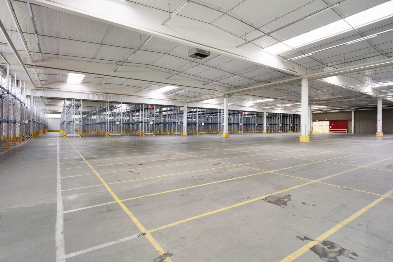 Stor, tom lagerhal med højt til loftet, gule parkeringspladser og flere lagerreoler langs venstre side. Kraftig loftsbelysning oplyser rummet.