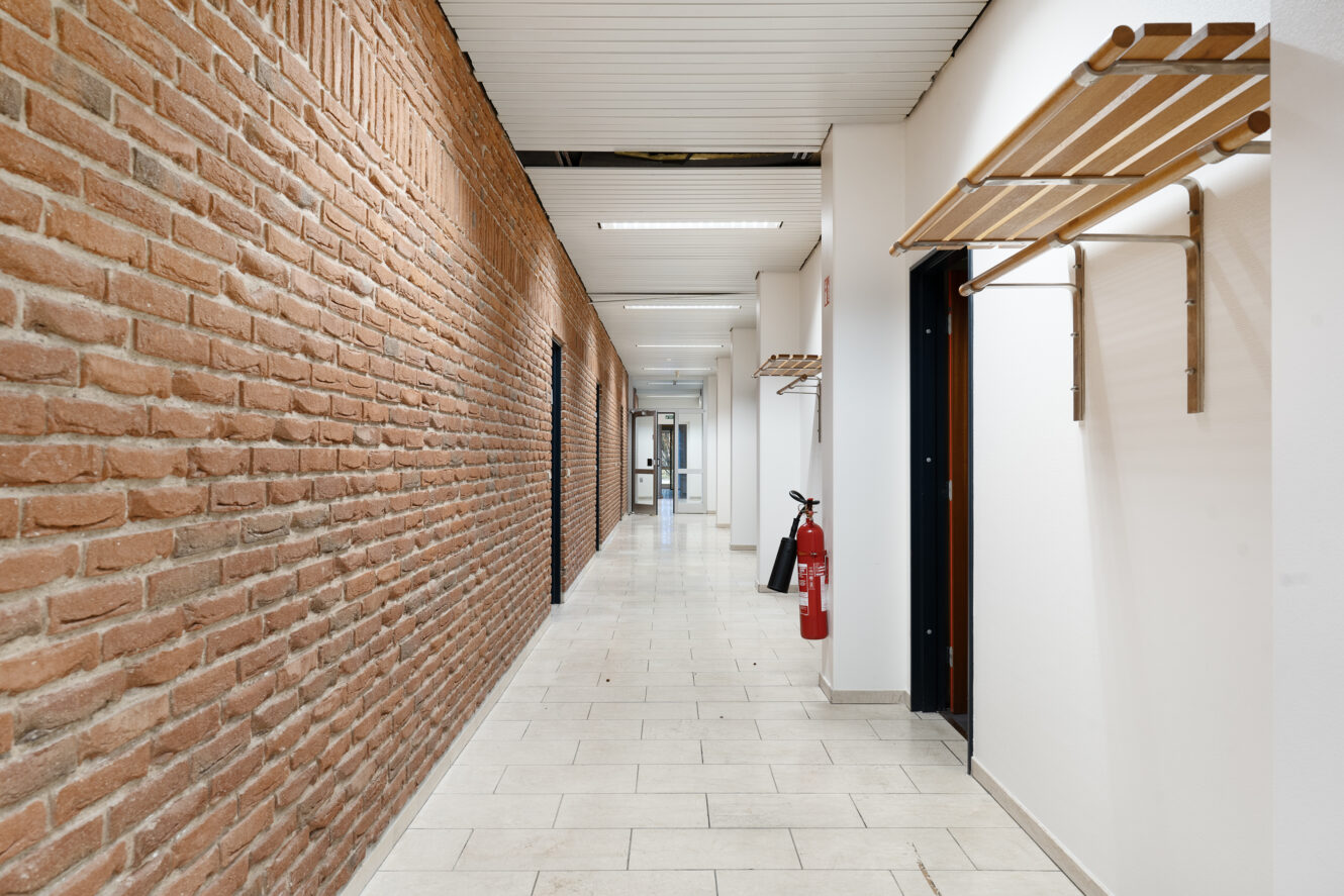 En gang med en murstensvæg til venstre, hvidt flisegulv, knagerækker til højre og en brandslukker ved siden af en døråbning.