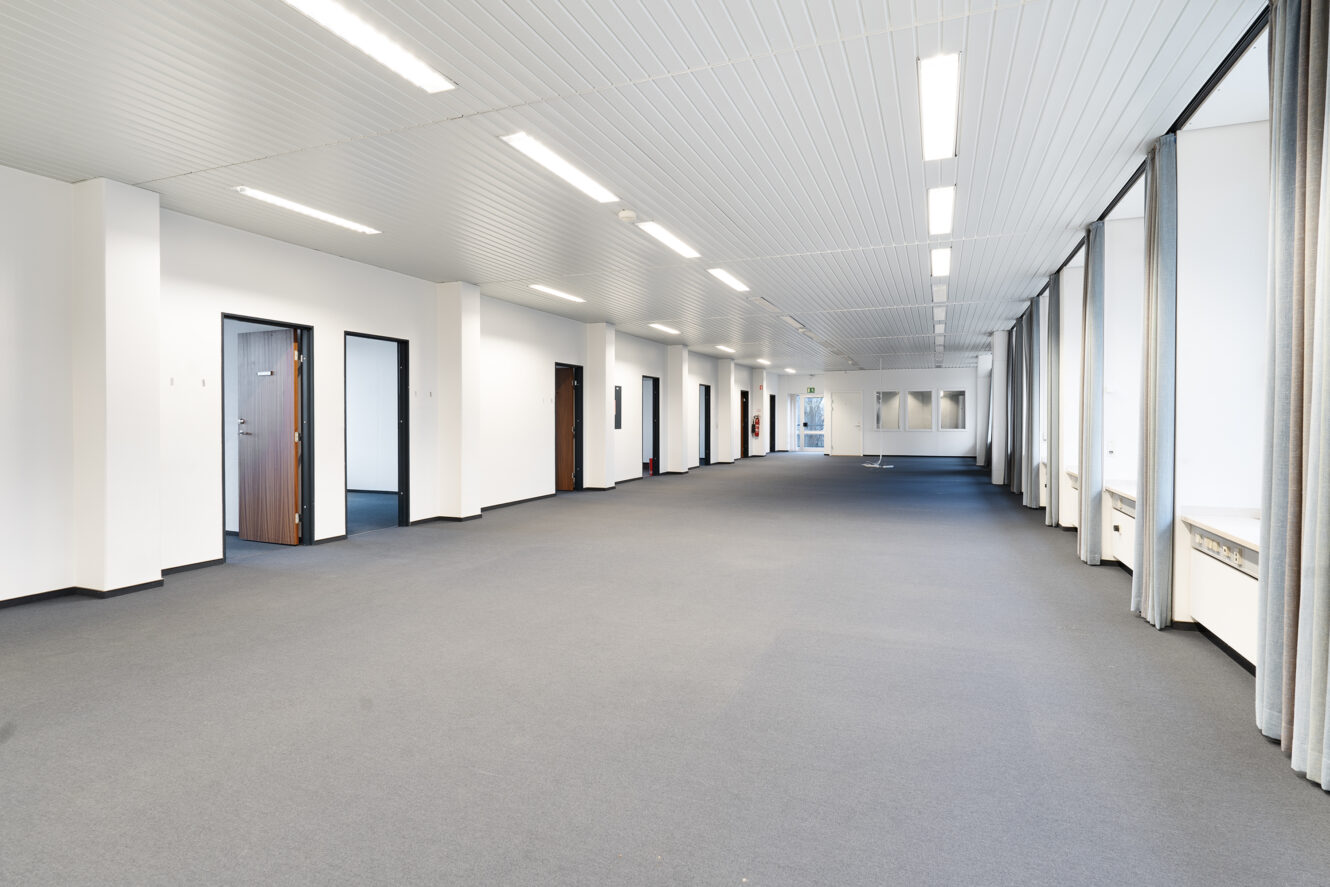 Et stort, tomt kontorlokale med gråt gulvtæppe, flere døråbninger til venstre, gulv-til-loft-vinduer med gardiner til højre og lysstofrør i loftet.