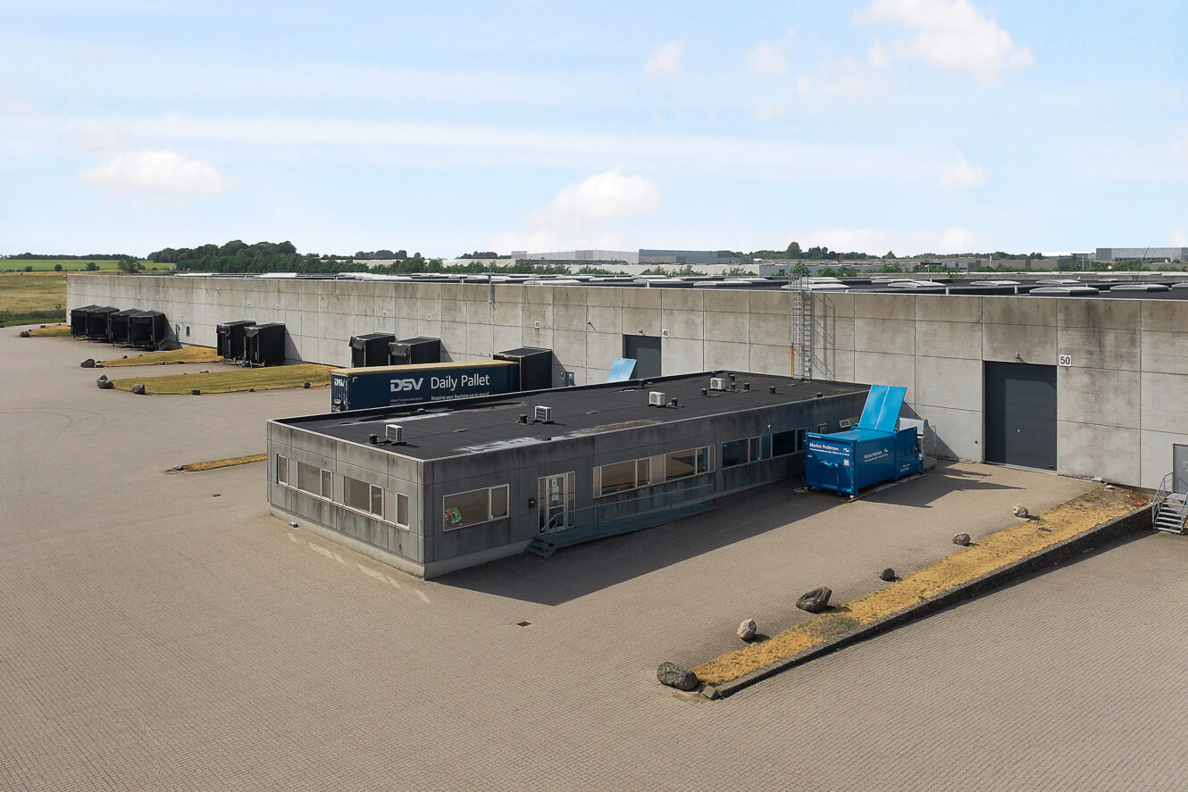 Et stort industrielt lagerkompleks med en kontorbygning med fladt tag i forgrunden, omgivet af asfalterede områder og læsseramper, under en delvist overskyet himmel.