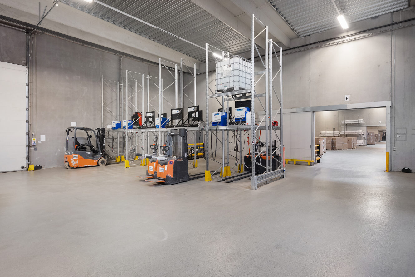 Gaffeltrucks og palleløftere er parkeret i nærheden af metalreoler med containere og udstyr i en ren, rummelig lagerhal med højt til loftet og betonvægge.