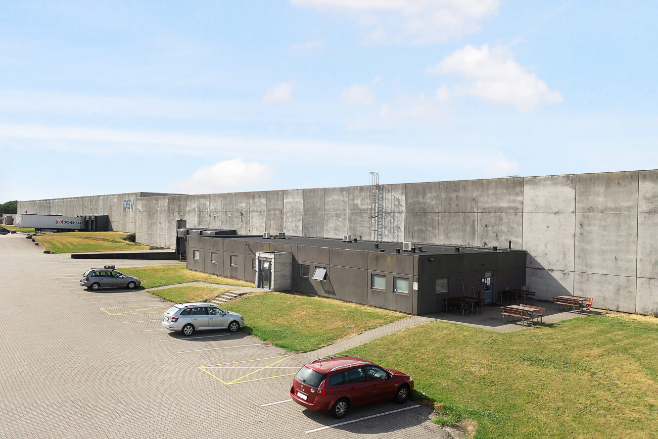 En enetages bygning med flere parkerede biler står foran en høj betonmur under en delvis overskyet himmel. Udendørs bænke er placeret på græsset ved siden af bygningen.