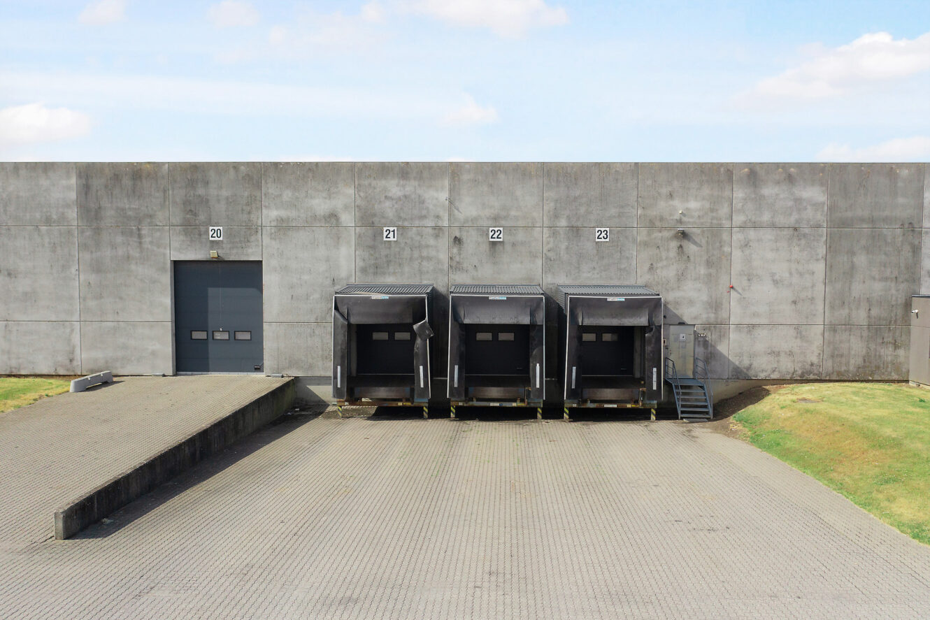 En læsserampe af beton med tre læsseramper, nummereret 21 til 23, og en lukket dør mærket 20 på venstre side af en stor, vinduesløs bygning.