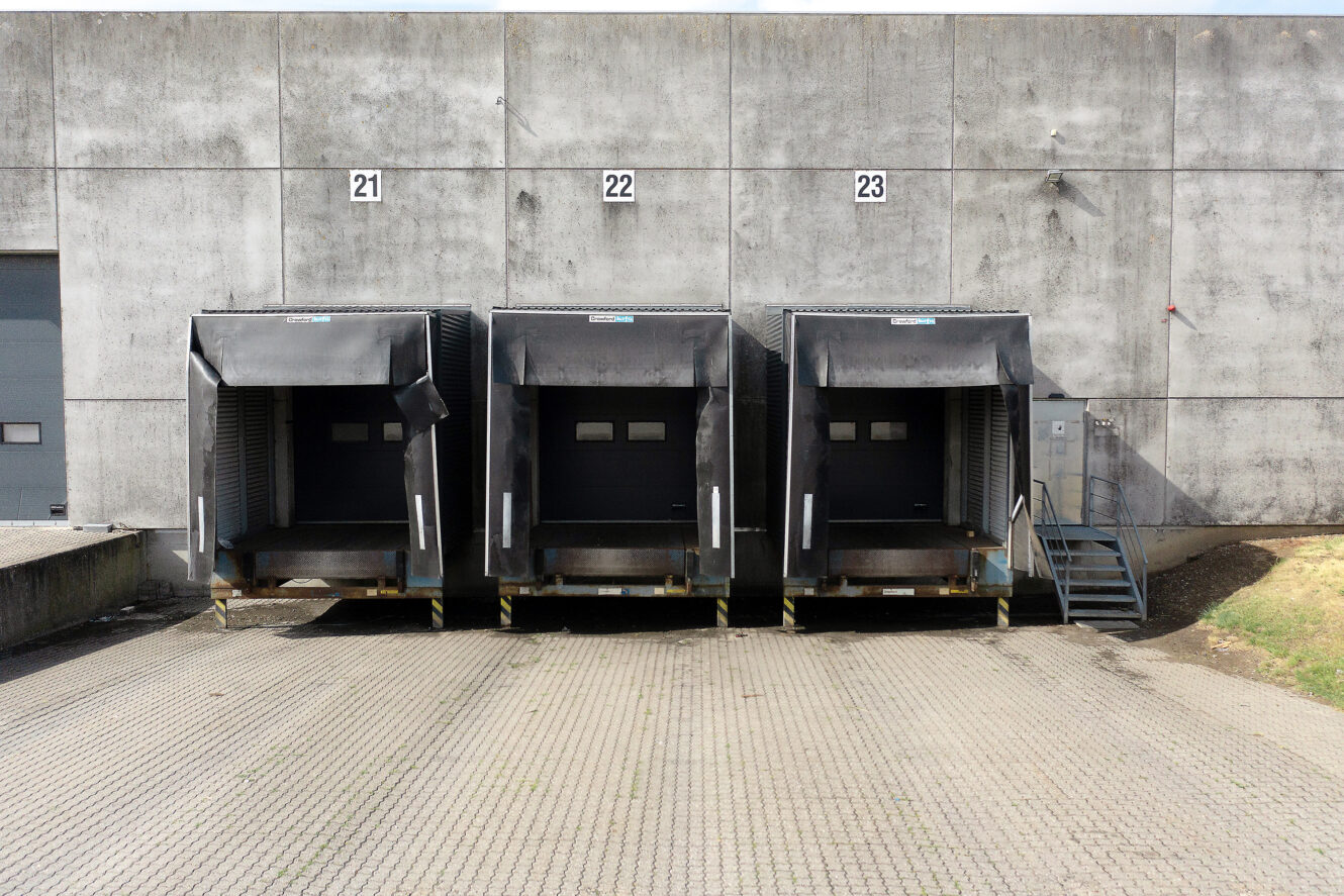 Tre læsseramper med numrene 21, 22 og 23 er stillet op mod en betonvæg i lagerhallen med lukkede dokdøre og en tilstødende metaltrappe.