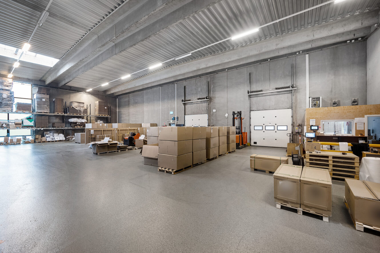 Rummeligt lagerinteriør med stablede papkasser på paller, højt til loftet, betonvægge og læsseramper i baggrunden.