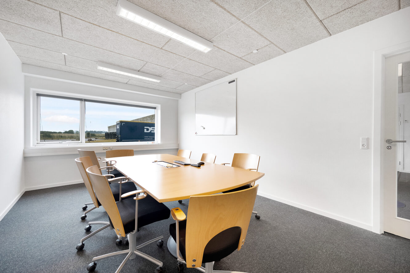 Et lille mødelokale med et rektangulært bord, seks stole, et whiteboard på væggen og et vindue, der lukker naturligt lys ind.