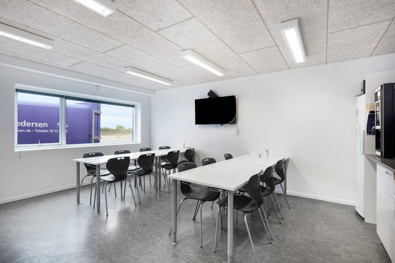 Et moderne, minimalistisk pauserum med to hvide borde, sorte stole, et væghængt tv, et vindue og ovenlys.