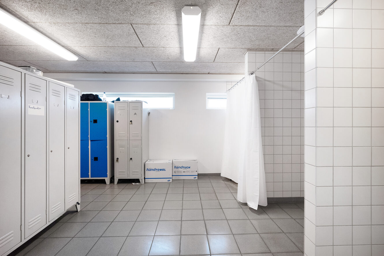 Et omklædningsrum med hvide og blå skabe, to bokse på gulvet, flisebelagte vægge og gulv og et bruseområde adskilt af et hvidt forhæng.