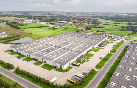 Luftfoto af et stort industrielt lagerkompleks med flere bygninger, læsseramper, parkerede lastbiler, grønne omgivelser og nærliggende veje.